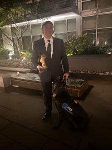 Chris Cadigan and Service Dog Brit stand outside holding up VETTY Statuette for 2021 in Suicide Prevention Category