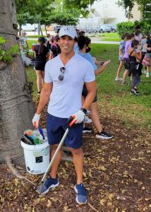 ALT="Dr. Amir Mohit-Kermani, a South Florida chiropractor, volunteers to pick up trash in a Miami Beach park and stands under a tree with a bucket of garbage"